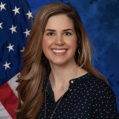 photo of a person with long blond hair and a navy shirt, there is a blue background with an American flag