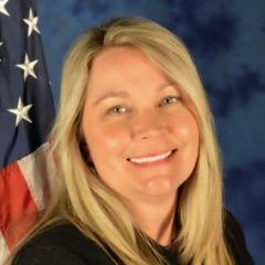 A woman sits in front of a blue background for her portrait. She is smiling and there is an American flag over her left shoulder.