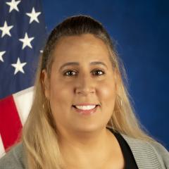 A woman sits in front of a blue background for her portrait. She is smiling and there is an American flag over her left shoulder.