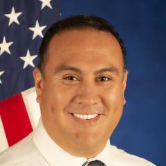 A man sits in front of a blue background for his portrait. He is smiling and there is an American flag over his left shoulder.