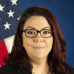 A woman sits in front of a blue background for her portrait. She is smiling and there is an American flag over her left shoulder.