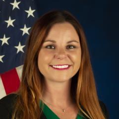 A woman sits in front of a blue background for her portrait. She is smiling and there is an American flag over her left shoulder.