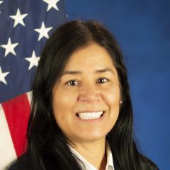 A woman sits in front of a blue background for her portrait. She is smiling and there is an American flag over her left shoulder.