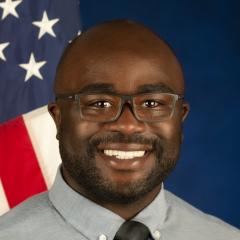 A man sits in front of a blue background for his portrait. He is smiling and there is an American flag over his left shoulder.