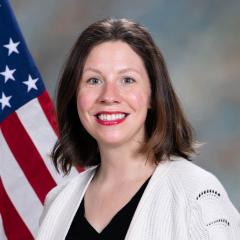 portrait of a person with shoulder-length brown hair, smiling at the camera, wearing a black shirt and a white cardigan; there is an American flag behind them