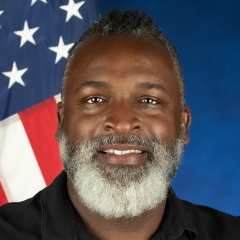 Photo of a man with a beard and short hair, smiling. He is sitting in front of a blue background and there is an American flag over his left shoulder.