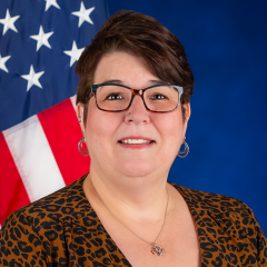 Portrait of a woman with short hair, wearing glasses and smiling. She is sitting in front of a blue background and the American flag is over her left shoulder.