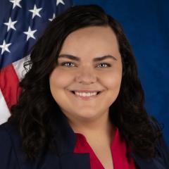 A woman sits in front of a blue background, with and American flag over her left shoulder, smiling for her professional portrait.