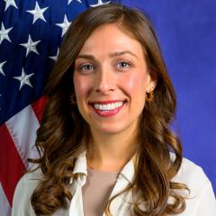 Woman with long brown hair seated in front of a U.S. Flag