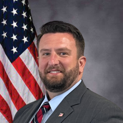 Male With Beard Wearing Gray Pinstripe Suit with tie standing in front of American Flag