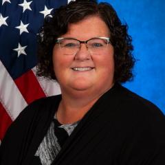 Angela is standing in front of an American flag dressed in a dark blazer with a marbled shirt. 