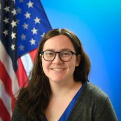A brunette woman wearing glasses posed in front of an American flag and blue background.