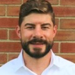 Man with dark hair and beard with light button up shirt.  Background is a brick wall.