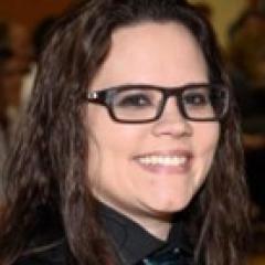 Woman with dark wavy hair, dark rimmed eyeglasses with dark colored shirt.