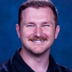 Man with short reddish brown hair, mustache and dark colored button down shirt.  Background of medium blue.