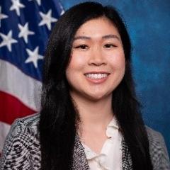 Young woman with long black hair, white blouse, black & white blazer.  Background is standard blue with American flag.