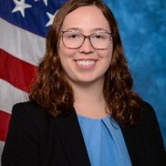 Young woman with dark hair, glasses, light blue blouse, dark blue or black blazer.  Background is standard blue with the American flag.