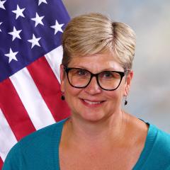 a person with short blonde hair wearing glasses sitting in front of US flag
