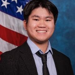 Young man with black hair, light blue shirt with dark blue/black tie and blazer.  Background is standard blue with American flag.