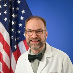 A man in a white lab coat posed in front of an American flag and blue background.