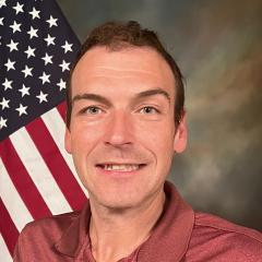 A person with short brown hair sitting in front of the US flag