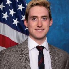 Young man with blond hair, white shirt, black tie and black and white tweed blazer.  Background standard blue with American flag