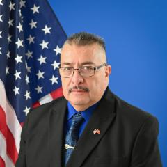 Man in a suit with an American flag and blue in the background.