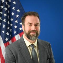 Man in a suit with a blue background and American flag.