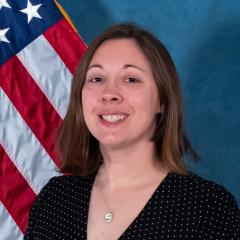 Woman with short brown hair, wearing a black dress with white dots, standing in front of an American flag.