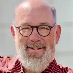 Close-up of an older man with glasses, a white beard, and an orange shirt smiling.