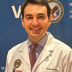 A smiling man in a white lab coat with a tie stands in front of a blue backdrop.