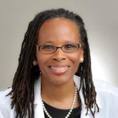 Woman with dreadlocks, glasses, white lab coat, and pearl necklace smiling.