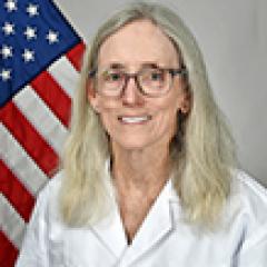 Woman in white lab coat smiling, with American flag in background.