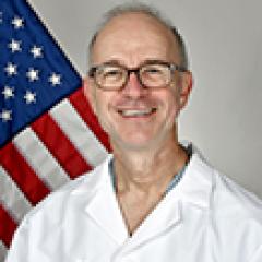Smiling man in a lab coat with an American flag behind him.