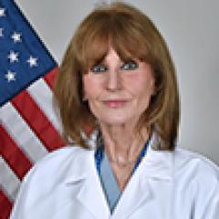 A smiling woman in a lab coat stands in front of an American flag.