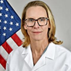 A woman in a lab coat stands in front of an American flag.