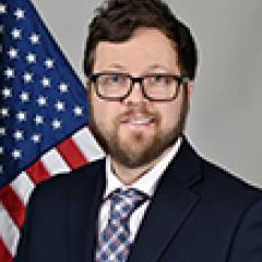 Man in a suit and tie smiling, with an American flag in the background.