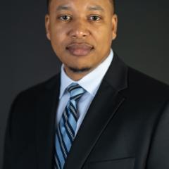 Man in a black suit and blue striped tie smiling against a black background.