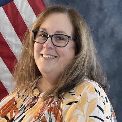 Woman with long hair wearing glasses and a patterned blouse, smiling in front of an American flag.