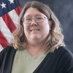 Portrait of a woman with glasses, wearing a green shirt and black cardigan, standing in front of a U.S. flag.