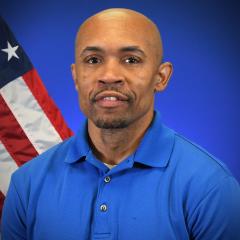 Tavares Moore in blue polo shirt stands in front of American flag.