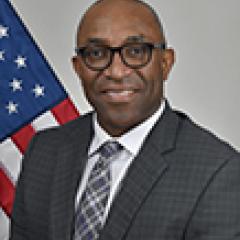 Smiling man in suit and tie standing in front of an American flag.