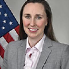 Woman in gray suit and pink shirt smiling with US flag behind her.