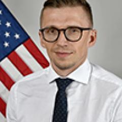 Man in a white dress shirt, black tie, and glasses stands in front of an American flag.