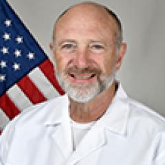 Bald man in a white lab coat smiles in front of an American flag.