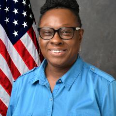 Woman with short hair and glasses, smiling in front of an American flag.