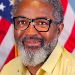 A smiling man with glasses and a yellow shirt stands in front of an American flag.