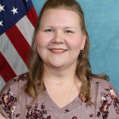 Woman with shoulder-length brown hair smiling with American flag in background.