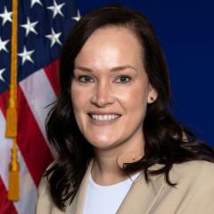 Woman with dark hair wearing a white shirt and beige blazer smiling in front of an American flag.