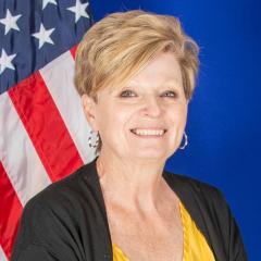 Smiling woman with short blonde hair, yellow top, and black cardigan in front of a flag.